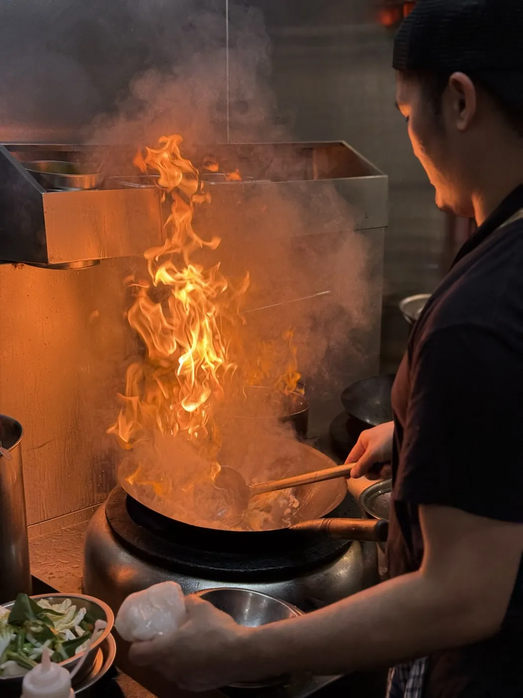 A cook tosses a wok over a tall flame in a busy kitchen.